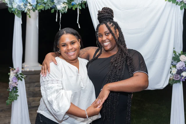 Two guests embracing and smiling beneath a floral arch